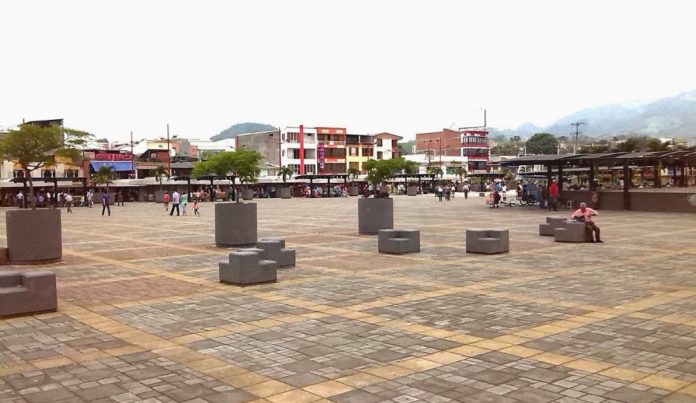 Una espaciosa plaza con suelo de baldosas, modernos asientos de bloques grises, árboles dispersos y gente paseando o sentada. Al fondo, edificios bajos de colores y la Biblioteca Pública Hugo Ángel Jaramillo, con montañas visibles a lo lejos.