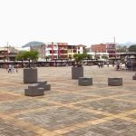 Una amplia plaza con modernos bancos grises, algunas personas sentadas y paseando, rodeada de coloridos edificios bajos y árboles, con la Biblioteca Pública dedicada a Hugo Ángel Jaramillo cerca y las montañas visibles al fondo.