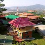 Vista aérea de una colorida urbanización en la ladera de una colina, con tejados rojos, verdes y marrones, rodeada de una exuberante vegetación; al fondo, bajo un cielo despejado, se ven las montañas y el paisaje urbano, en el que destaca la Biblioteca Pública.