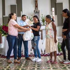 Cinco personas permanecen juntas en el interior, sonriendo mientras una mujer entrega una bolsa blanca a otra. Detrás de ellas, flores, velas y grandes letras blancas crean un ambiente festivo que recuerda a las celebraciones de las estaciones Balboa.