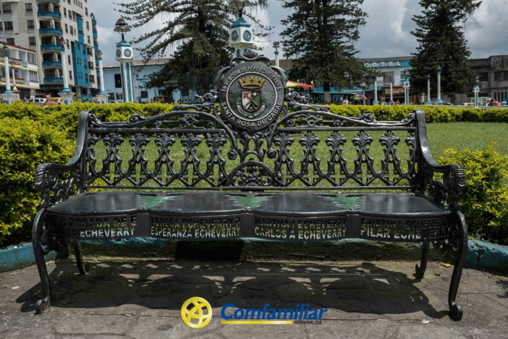 Cerca de la Biblioteca Pública, rodeado de vegetación, edificios y altos árboles, se encuentra un banco decorativo de metal negro con diseños ornamentales y un escudo en el centro. En su asiento hay nombres grabados, entre ellos el de Jaime Fernández Botero.