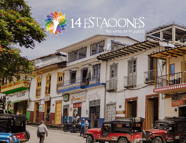 Escena callejera en la colorida Apía, Risaralda, con balcones tradicionales, gente caminando y Jeeps estacionados. El texto 14 Estaciones: Nos vemos en mi pueblo con un vibrante logotipo aparece en la parte superior.