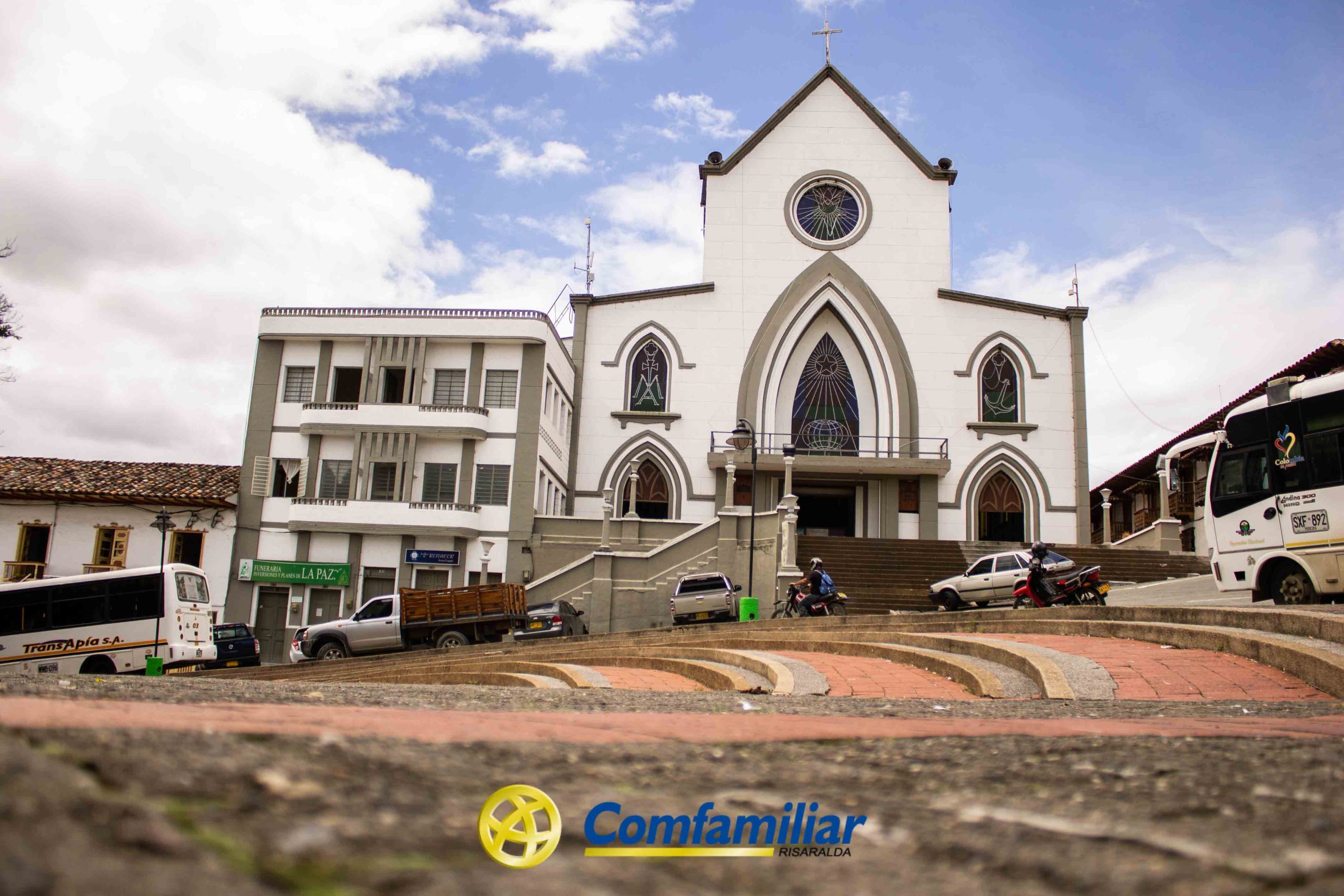 Una iglesia blanca con ventanas arqueadas y una cruz en lo alto se alza junto al edificio Municipal Isaías Naranjo. Al frente se ven vehículos y personas. El logotipo de Comfamiliar Risaralda aparece en la parte inferior de la imagen.