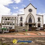 Una iglesia blanca con una gran ventana arqueada se alza detrás de una plaza escalonada. Delante, varios vehículos y personas. A la izquierda se ve el edificio de la Biblioteca Pública Isaías Naranjo. El logotipo de Comfamiliar Risaralda aparece en la parte inferior de la imagen.