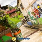 Un colorido callejón con murales de pájaros y flores, plantas en macetas y un letrero de madera que dice Vilo de los Cascacones. En la parte inferior aparecen los logotipos de la Biblioteca Pública Municipal Isaías Naranjo y Comfamiliar Risaralda.