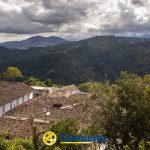 Vista panorámica de tejados de tejas en un pueblo rodeado de exuberantes colinas verdes bajo un cielo nublado, con montañas a lo lejos. El logotipo de la Biblioteca Pública Isaías Naranjo Confamiliar aparece en la parte inferior central de la imagen.