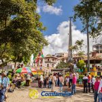 Una animada plaza con gente reunida, sombrillas de colores, árboles y varios edificios. Una estatua y un monumento alto -cerca de la Biblioteca Pública Isaías Naranjo- son visibles en el centro. El logotipo de Comfamiliar aparece en la parte inferior de la imagen.