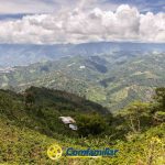 Amplia vista de exuberantes montañas verdes y valles bajo un cielo parcialmente nublado, con casas dispersas y tierras de cultivo. La Biblioteca Pública y el logotipo de Confamiliar aparecen en la parte inferior central de la imagen.