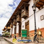 Una estrecha calle adoquinada con edificios de estilo colonial con balcones de madera. Una bicicleta amarilla con cestas está aparcada junto a una farola, cerca de la Biblioteca Pública Isaías Naranjo. Unas macetas decoran la escena. En la parte inferior se ve el logotipo de Comfamiliar.