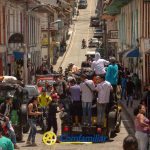 Multitudinaria escena callejera en un colorido pueblo cerca de la Biblioteca Pública Municipal. Muchas personas se reúnen alrededor de vehículos, algunos montados en camiones. Edificios con balcones se alinean a ambos lados de la accidentada calle. En el bullicioso ambiente aparece el logotipo de Comfamiliar en la parte inferior.