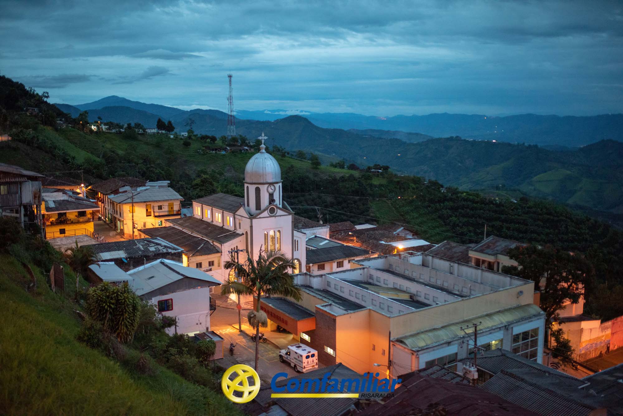 Una pequeña ciudad con una iglesia y una cúpula blancas está enclavada en las verdes colinas de Risaralda al atardecer, con cálidas farolas que iluminan los edificios. Al fondo se ven montañas y un cielo nublado. El logotipo de Comfamiliar Risaralda aparece en la parte inferior.