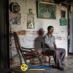 Un hombre se sienta en una silla dentro de una habitación rústica con paredes desgastadas, fotos familiares y una mecedora. La luz de la izquierda resalta su expresión contemplativa. El logotipo de Comfamiliar y la Biblioteca Pública Municipal Juan XXIII aparecen en la parte inferior.