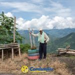 Un hombre mayor se encuentra al aire libre en un camino de montaña junto a una jardinera de neumáticos pintados, bancos de madera y exuberantes colinas verdes bajo un cielo nublado, representando a la Biblioteca Pública Hugo Ángel Jaramillo. El logotipo de Confamiliar aparece en la parte inferior de la imagen.