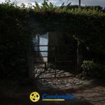 Una verja metálica situada en un seto conduce a un camino empedrado, rodeado de densa vegetación. Una luz brillante atraviesa la verja en dirección a la Biblioteca Pública Municipal, y en la parte inferior de la imagen aparece el logotipo de Comfamiliar.