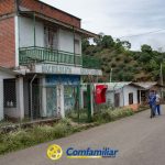 Un hombre y un niño caminan por una calle tranquila junto al edificio de la escuela Municipal Juan XXIII, con paredes pintadas y un balcón verde, en una zona rural rodeada de árboles. El logotipo de Comfamiliar Risaralda aparece en la parte inferior de la imagen.