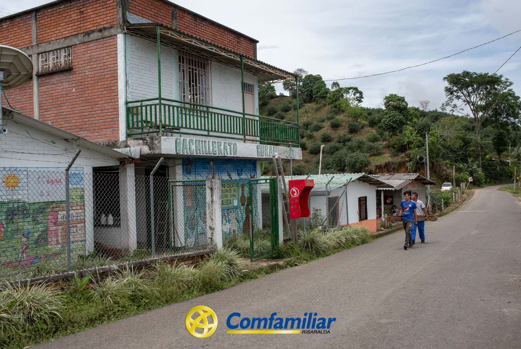 Un hombre y un niño caminan por una calle tranquila junto al edificio de la escuela Municipal Juan XXIII, con paredes pintadas y un balcón verde, en una zona rural rodeada de árboles. El logotipo de Comfamiliar Risaralda aparece en la parte inferior de la imagen.