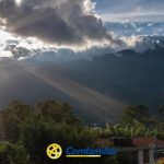 La luz del sol se cuela entre las nubes sobre verdes montañas y árboles, con algunos tejados visibles abajo, cerca de la Biblioteca Pública Municipal. El logotipo y el nombre de Comfamiliar aparecen en la parte inferior de la imagen.