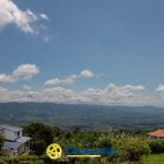 Paisaje rural con cielo azul, nubes blancas, montañas lejanas y dos casas rodeadas de vegetación. En la parte inferior se superpone el logotipo de la Biblioteca Pública Confamiliar.