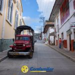 Un Jeep rojo de época está aparcado en una tranquila calle bordeada de coloridos edificios de estilo colonial, cerca de la Biblioteca Pública Municipal. Algunas personas caminan por la acera. El logotipo de Comfamiliar aparece en la parte inferior de la imagen.