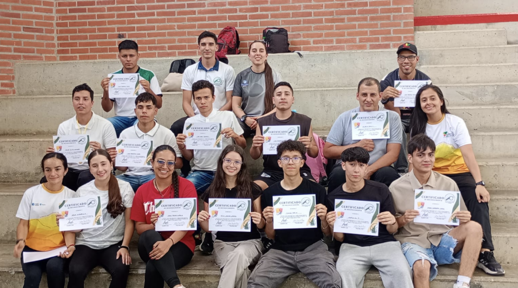 Un grupo de personas sentadas en gradas de cemento en Risaralda, sonríen y sostienen certificados. La mayoría son adultos jóvenes, vestidos informalmente, celebrando su reciente logro en el PAD de arbitraje de voleibol.