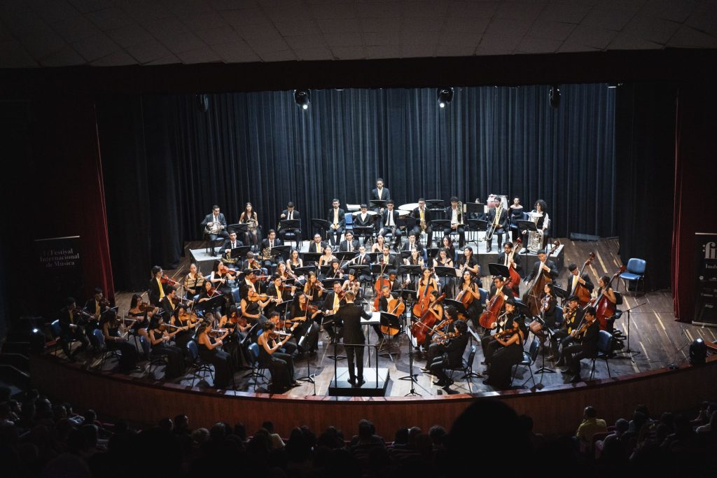 Una gran orquesta, como la Orquesta Sinfónica Nacional de Colombia, actúa en el escenario bajo luces brillantes, con músicos que tocan varios instrumentos mientras un director los dirige ante un público sentado en un gran concierto en la sala de conciertos.