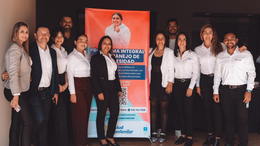 Un grupo de doce personas vestidas con traje de negocios sonríen delante de una pancarta que promociona el programa de salud "ruta de obesidad", dentro de una habitación bien iluminada. En la pancarta aparece una mujer con bata blanca y texto en español.