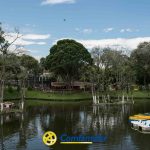 Una tranquila escena de parque muestra un lago con botes de remos, árboles a lo largo de la orilla y algunos pequeños muelles. El logotipo de Comfamiliar Risaralda y la Biblioteca Pública aparecen en la parte inferior central de la imagen.