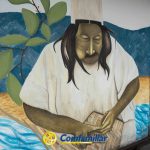 Un mural en Dosquebradas representa a un indígena con vestimenta tradicional, concentrado mientras teje con sus manos. El fondo muestra hojas verdes, montañas azules y agua corriente. El logotipo de Comfamiliar aparece en la parte inferior.