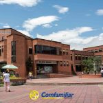 La gente camina en una plaza frente a la gran Biblioteca Pública de ladrillo rotulada BIBLIOTECA MUNICIPAL e ICAM, con palmeras y el logo de Comfamiliar Risaralda en la parte inferior. La escena está ambientada en Dosquebradas bajo un cielo parcialmente nublado.