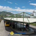 Un autobús amarillo está aparcado en un andén cubierto de la estación de Dosquebradas, rodeado de barandillas verdes y colinas. El logotipo de Comfamiliar Risaralda y el letrero de la Biblioteca Pública son visibles bajo un cielo parcialmente nublado.