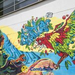 Colorido mural en la pared de un edificio de Dosquebradas con un vibrante loro en vuelo, grafitis abstractos, un rostro humano con tocado y remolinos amarillos. El logotipo de Comfamiliar aparece en la parte inferior, cerca de la Biblioteca Pública.