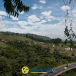 Una carretera en curva serpentea entre frondosas colinas verdes y casas dispersas bajo un cielo parcialmente nublado. En primer plano, el logotipo de Comfamiliar Dosquebradas aparece en la parte inferior, destacando su conexión con la cercana Biblioteca Pública.