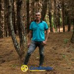 Un hombre vestido con camisa azul, vaqueros y botas de goma se encuentra entre altos pinos en un bosque cerca de Guática. El suelo está cubierto de agujas de pino. El logotipo de Comfamiliar Guática aparece en la parte inferior de la imagen.