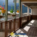 Un pasillo de madera con macetas de flores colgando del techo en la Biblioteca Pública Comfamiliar Guática. El pasillo da a tejados y montañas lejanas, con el suelo desgastado y la luz del sol proyectando sombras. El logotipo de Comfamiliar es visible en la parte inferior.