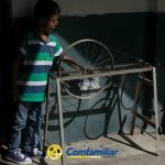 Un joven con camisa de rayas está de pie en el interior de la Biblioteca Pública, tocando una rueda de bicicleta montada en un bastidor metálico. La luz del sol proyecta sombras en la pared. El logotipo de Comfamiliar Guática aparece en la parte inferior de la imagen.