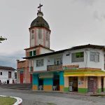 Una iglesia con una cúpula y una cruz se alza detrás de la Biblioteca Pública y un edificio de dos plantas pintado de amarillo, verde y blanco en una esquina de una tranquila plaza, con el cielo nublado.