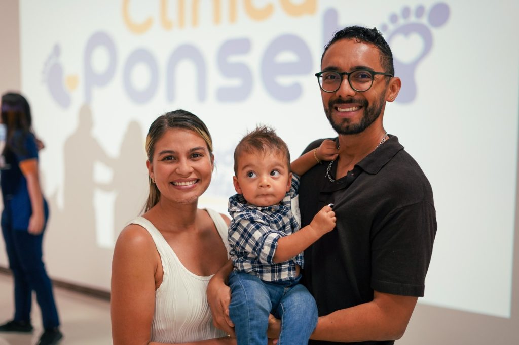 Una pareja sonriente sostiene a un bebé con pie equino varo delante de una pared con el logotipo de la Clínica Ponseti, que destaca la atención especializada que reciben. La mujer lleva un top blanco, el hombre gafas y camisa negra, y el bebé una camisa de cuadros.