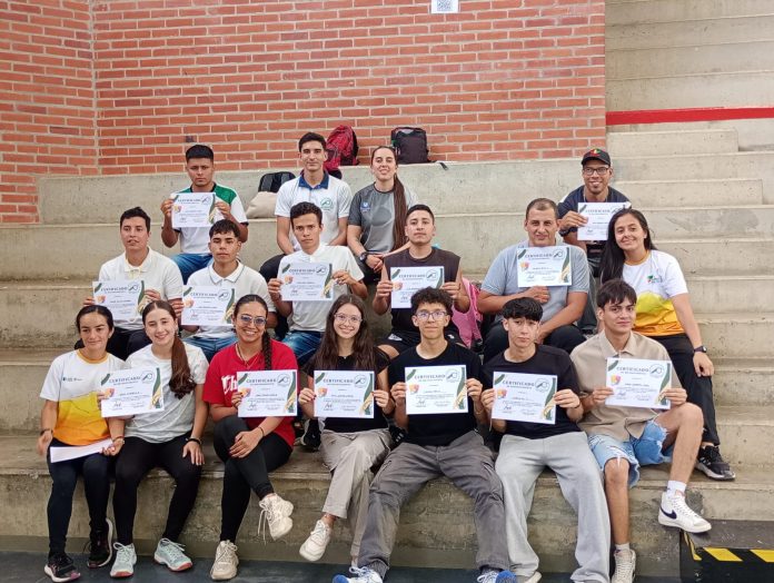 Un grupo de personas sentadas en gradas en Risaralda, sosteniendo certificados de un evento PAD de arbitraje de voleibol y sonriendo a la cámara. El fondo muestra una pared de ladrillo y escalones de concreto.
