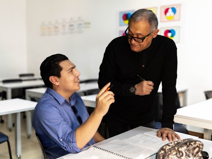 Un estudiante de educación básica está sentado en un pupitre con un cuaderno abierto, sonriendo y hablando con un profesor de pie en el aula. El profesor, que sostiene un bolígrafo y le devuelve la sonrisa, está rodeado de gráficos de colores en la pared detrás de ellos.
