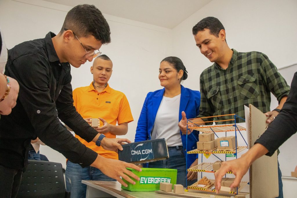 Cinco personas de pie alrededor de una mesa con maquetas de contenedores de transporte y un almacén, sonríen y participan en una actividad colaborativa, mostrando el aprendizaje y el trabajo en equipo a través de un proyecto de Educación Comfamiliar o Microcredenciales.