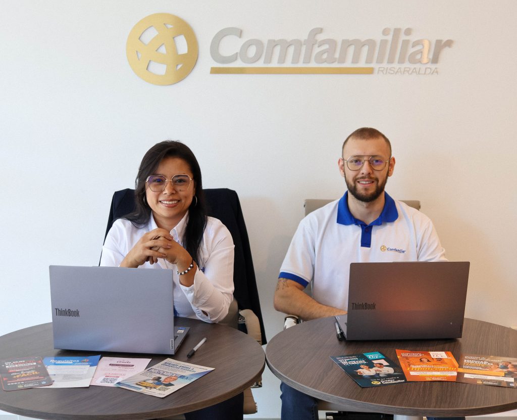 Dos personas sentadas en mesas redondas separadas con ordenadores portátiles y folletos, sonríen a la cámara. Detrás de ellos, el logotipo de Comfamiliar Risaralda marca este acogedor punto de atención de servicios.