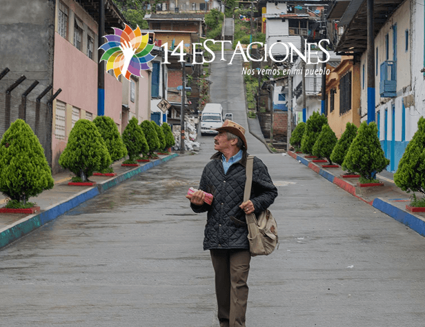 Un hombre con sombrero y bolso camina por una calle tranquila y arbolada de La Celia, Risaralda. El logo de 14 Estaciones y la frase Nos vemos en mi pueblo aparecen en la parte superior de la imagen.