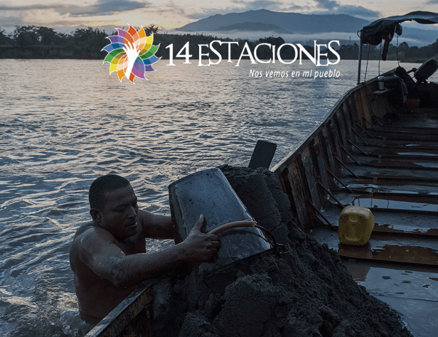 Un hombre se encuentra en un río al atardecer cerca de La Virginia, Risaralda, subiendo un cubo de arena a una barca de madera. El logotipo de "14 Estaciones" y la frase "Nos vemos en mi pueblo" aparecen en la parte superior de la imagen.