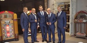 Cinco hombres vestidos con trajes azules sonríen en una sala de temática retro en la que se exponen tocadiscos y radios de época que capturan el espíritu de la Serenata Consotá y Los Ayer's.