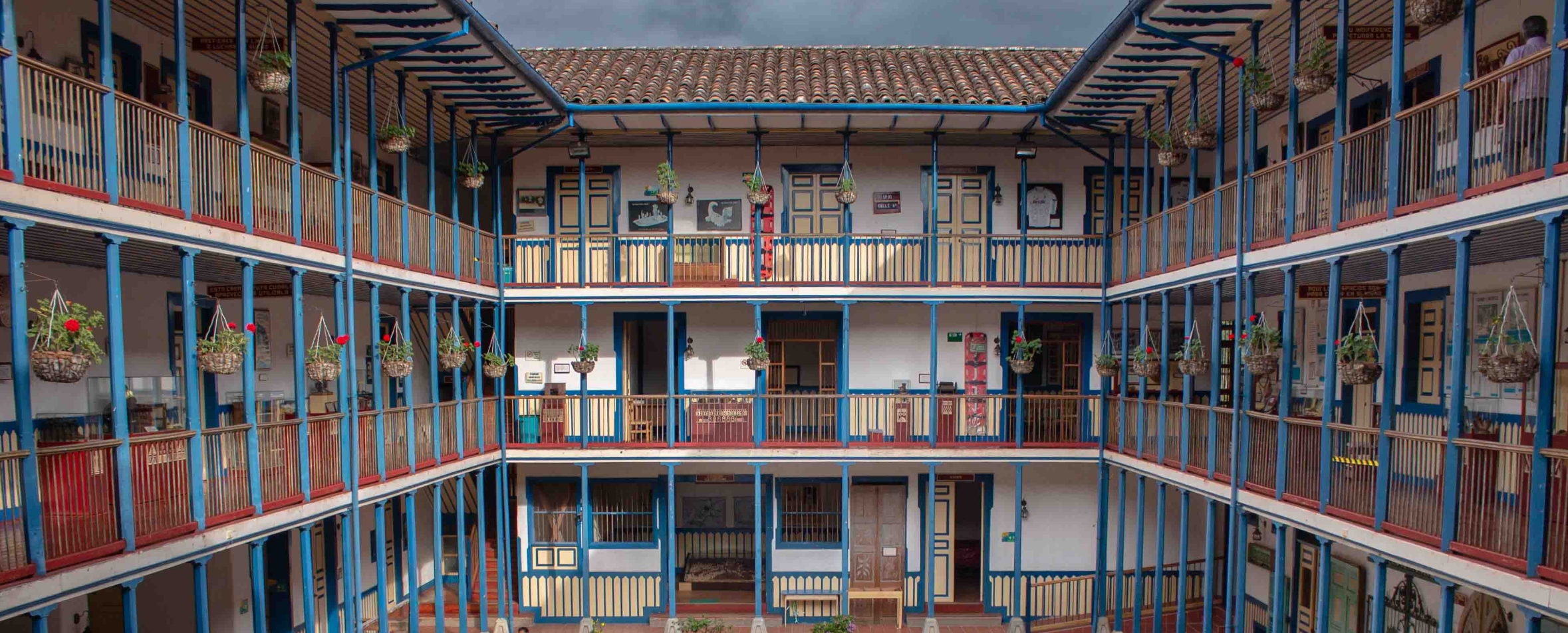Un tradicional edificio colombiano con patio, sede de la Biblioteca Pública Municipal León de Greiff, con tres coloridas plantas, barandillas de madera, cestas de flores colgantes y puertas que dan a un espacio central abierto bajo un cielo nublado.
