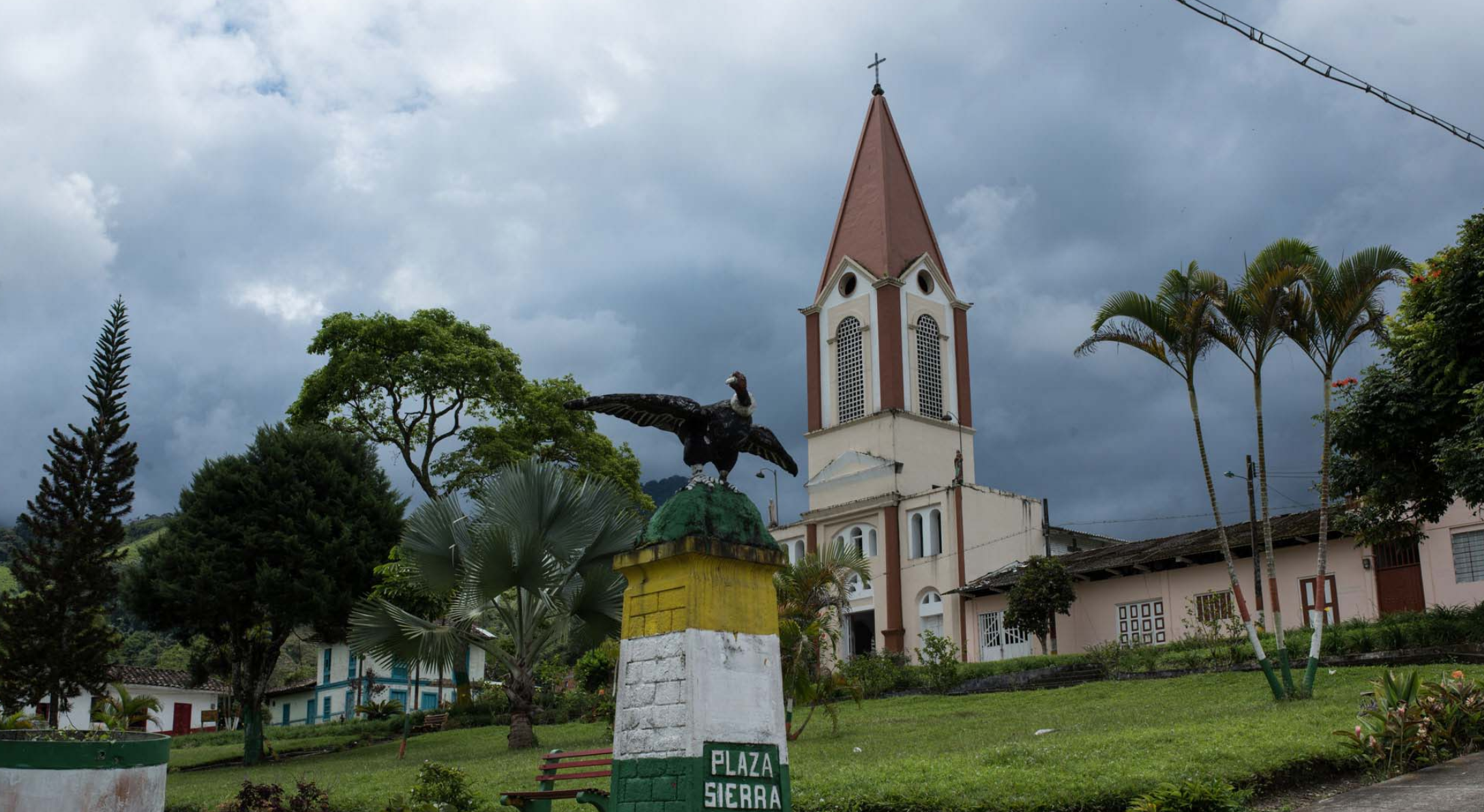 Una plaza en Mistrató con una estatua de un gran pájaro sobre un pedestal llamada Plaza Sierra, rodeada de vegetación, palmeras, la Biblioteca Pública y una iglesia con un alto campanario bajo un cielo nublado.