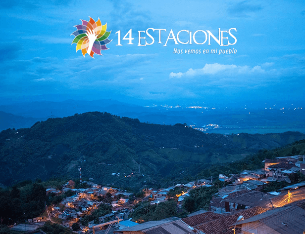 Vista panorámica de una pequeña ciudad enclavada en verdes colinas al atardecer cerca de 14 estaciones Pereira, con tejados en primer plano y montañas detrás. El texto 14 Estaciones: Nos vemos en mi pueblo y un logotipo multicolor sobre el cielo.