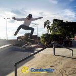 Un skater realiza un truco en el aire sobre una barandilla en un parque de skate al aire libre a la luz del día, con árboles, gente y el logotipo de Comfamiliar visible cerca de la Biblioteca Pública en primer plano.