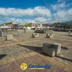 Una plaza amplia y abierta con bancos de hormigón y jardineras de piedra bajo un cielo azul salpicado de nubes. Edificios, carpas y palmeras en el fondo, cerca de la Biblioteca Pública Hugo Ángel Jaramillo. En la parte inferior aparece el logotipo de "Comfamiliar Risaralda".