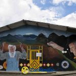 Un colorido mural sobre un edificio, obra de Hugo Ángel Jaramillo, muestra a dos ancianos, uno de ellos con bastón, rodeados de montañas, flores y un pájaro. Debajo aparecen diseños geométricos con la Biblioteca Pública Comfamiliar en la parte inferior central.